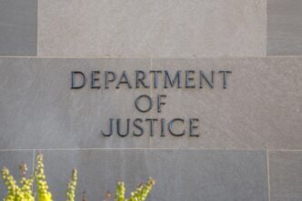 Close-up of raised, dark metallic letters reading "DEPARTMENT OF JUSTICE" on a beige stone wall.