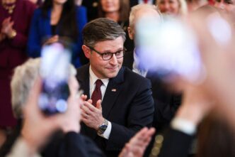 Speaker Johnson, wearing glasses and a suit, smiles while clapping at a White House event.