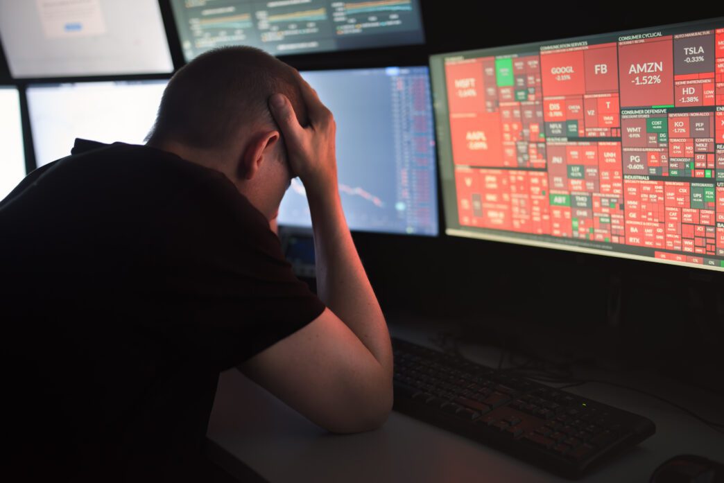 Man with his head in his hands, slumped over a keyboard, watching a red screen showing a massive stock market plunge.