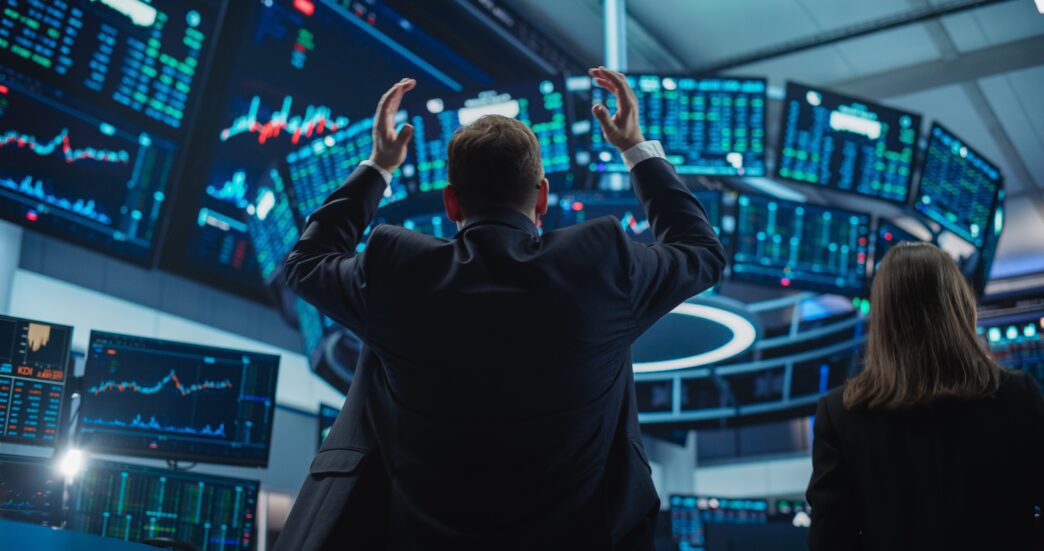 Depressed stock trader in a suit with hands raised in frustration, surrounded by multiple monitors displaying negative financial data.