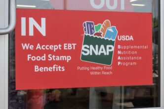 Red sign on a store window stating "We Accept EBT Food Stamp Benefits" and featuring the USDA SNAP logo.