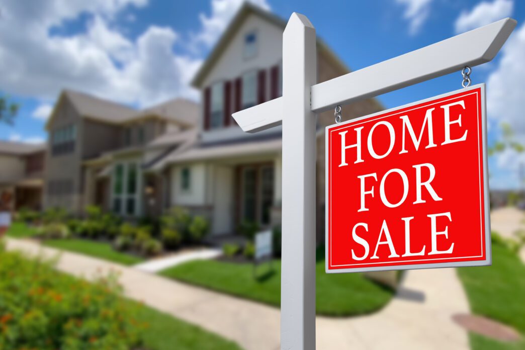 Red "HOME FOR SALE" sign in front of a blurry, two-story suburban house on a sunny day.