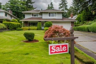 A wooden post with a white and red "Sale" sign is placed on the lawn in front of a large suburban house.