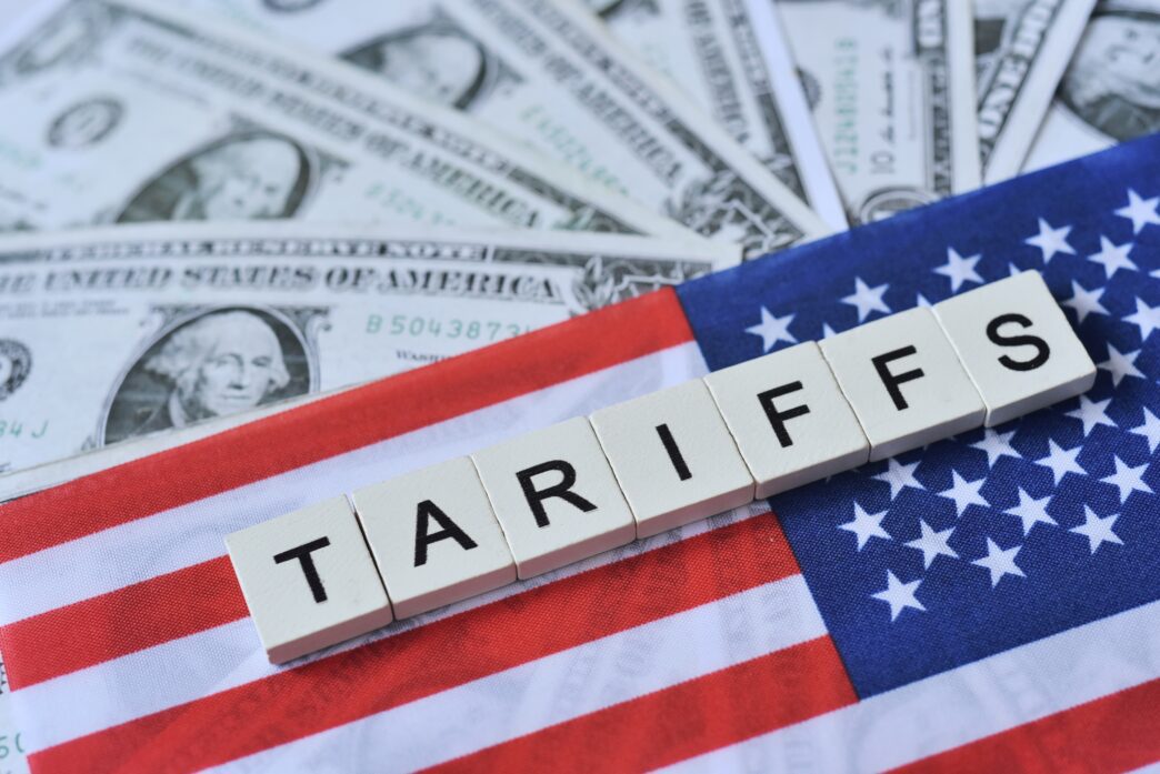 Wooden scrabble tiles spelling "TARIFFS" placed horizontally across a folded US flag, with US dollar bills fanned out in the background.