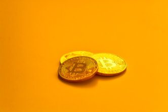 Three physical gold Bitcoin coins lying flat and overlapping on a brightly lit, solid orange background.
