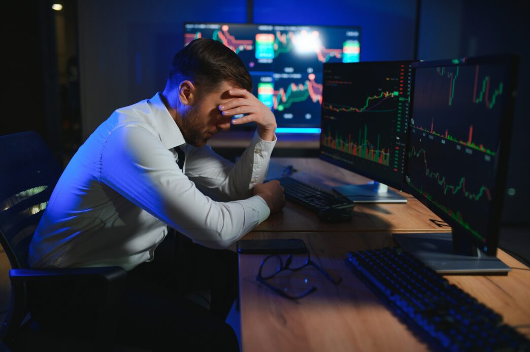 Stressed crypto trader with his head in his hand, facing multiple volatile market monitors.