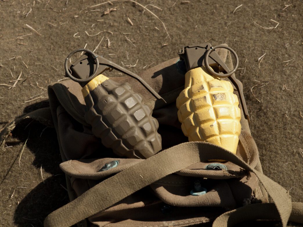 Two pineapple-style hand grenades, one dark green and one yellow, resting on a soldier's canvas bag.