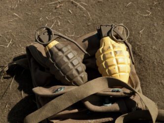 Two pineapple-style hand grenades, one dark green and one yellow, resting on a soldier's canvas bag.