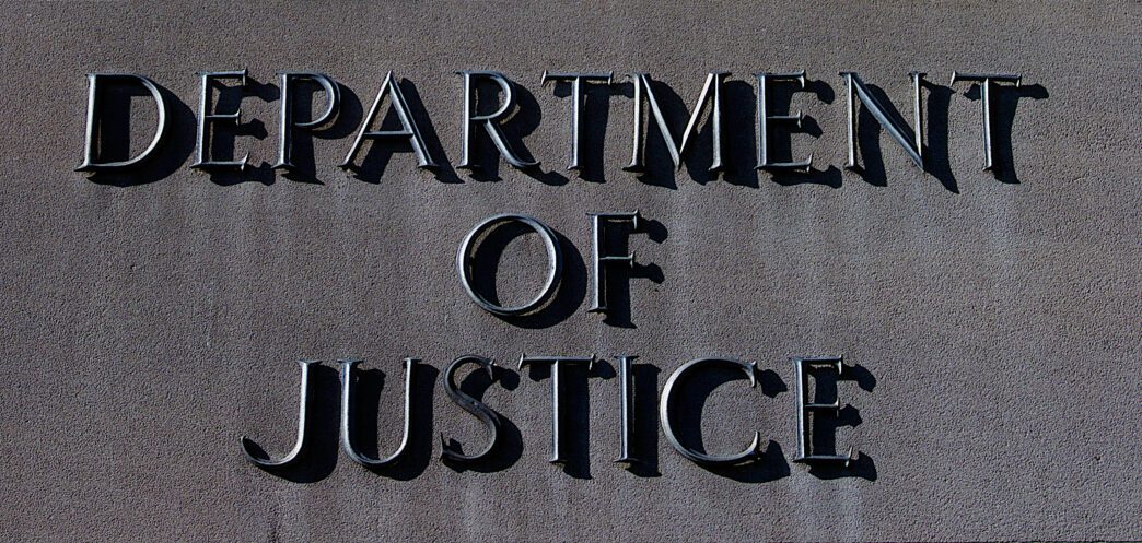 Close-up of the large, raised, metallic letters reading "DEPARTMENT OF JUSTICE" on a gray stone wall.