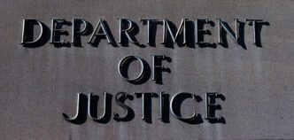 Close-up of the large, raised, metallic letters reading "DEPARTMENT OF JUSTICE" on a gray stone wall.