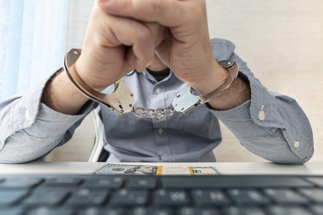 Man handcuffed at his desk, with his hands resting over a hundred-dollar bill and a keyboard.