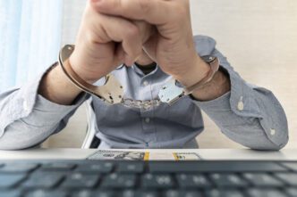 Man handcuffed at his desk, with his hands resting over a hundred-dollar bill and a keyboard.