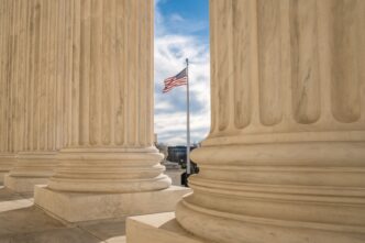 View of the US flag between two large, fluted columns of the Supreme Court building.