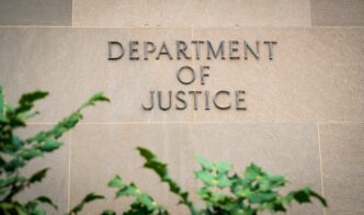 Raised letters reading "DEPARTMENT OF JUSTICE" on a beige stone wall, partially framed by green plants.