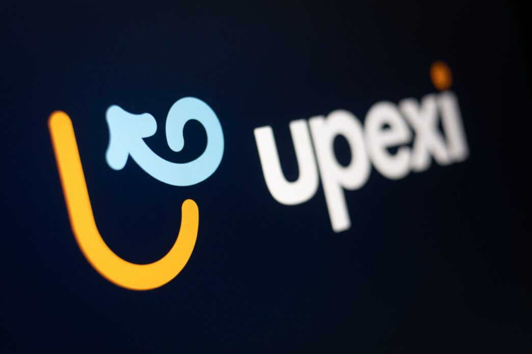 Close-up of the Upexi logo on a dark screen, showing the yellow U-shape and blue arrow-like swirl preceding the white text "upexi."