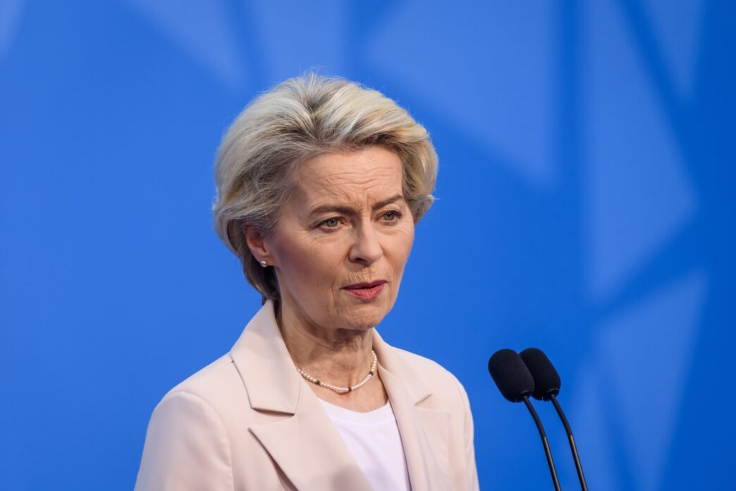 Close-up of Ursula von der Leyen, President of the European Commission, speaking at a podium against a solid blue background.