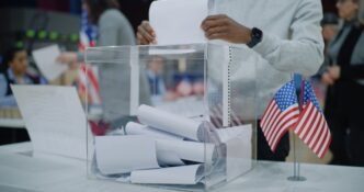 A voter drops a folded paper ballot into a transparent ballot box at a US polling station.