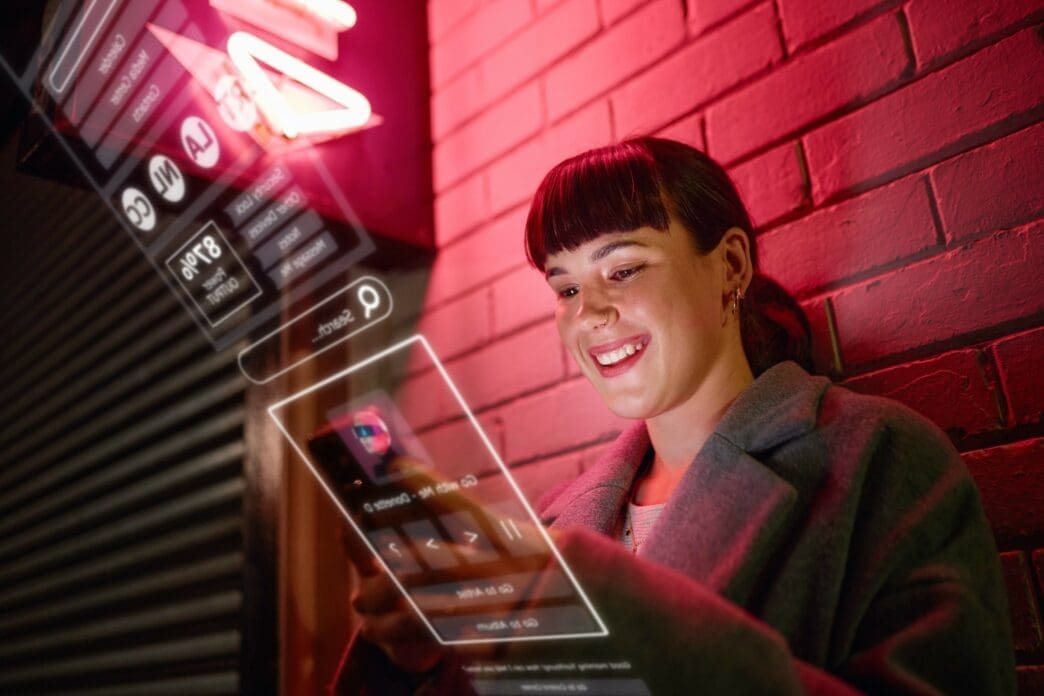 Smiling woman using a translucent phone with digital interface projections against a red brick wall at night.