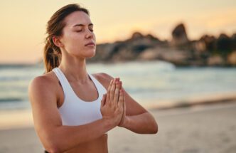 Woman in a white sports bra practicing yoga meditation on a beach at sunset with hands in Namaste.