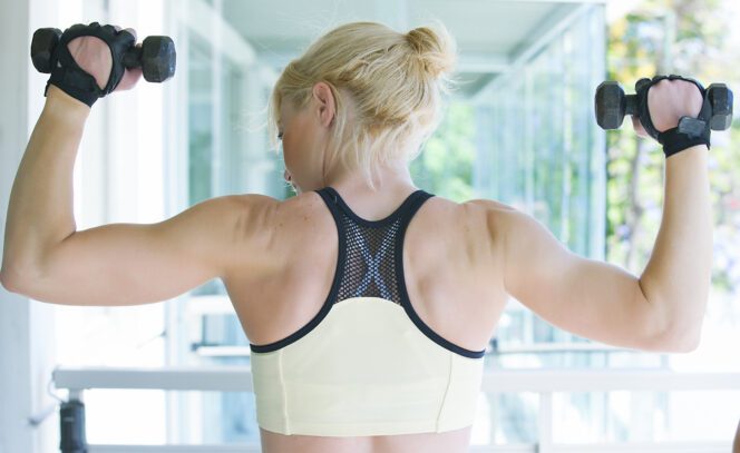Rear view of a woman lifting dumbbells, showcasing muscular shoulders and back during a workout.