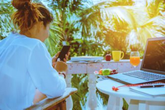Woman using a phone and laptop outdoors on a tropical balcony, enjoying a workation.