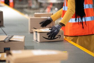 A logistics worker in a yellow shirt and orange vest places an Amazon package onto a conveyor belt for sorting.