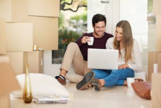 Happy young couple sitting on the floor using a laptop, surrounded by moving boxes.