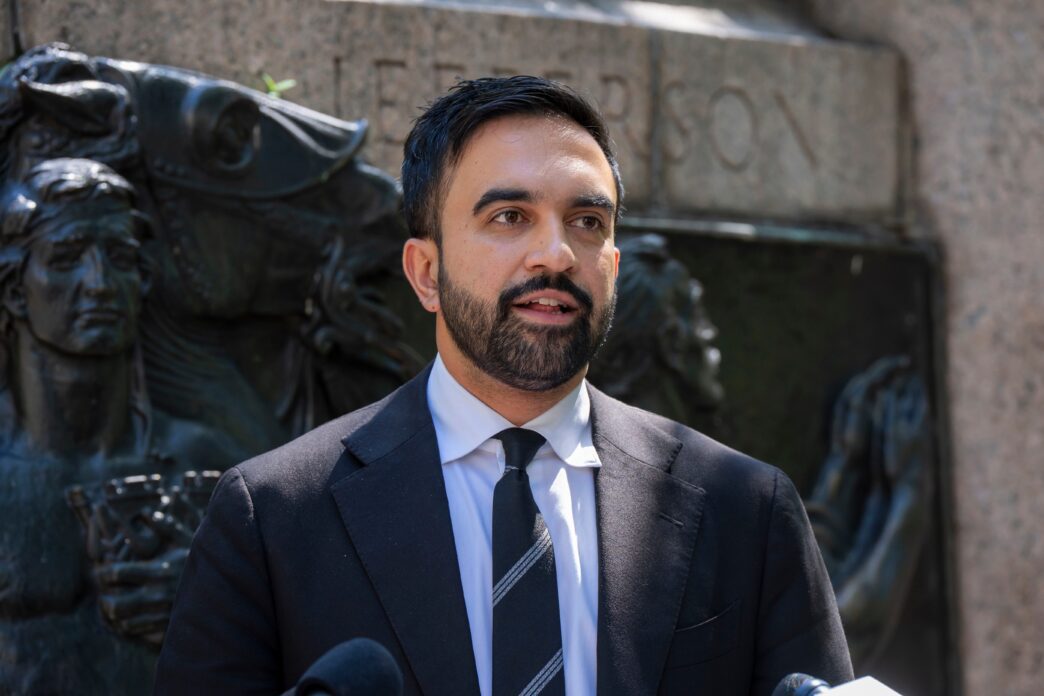 Zohran Mamdani, a man with a beard, speaks at a press briefing in a suit and tie in front of a stone monument.