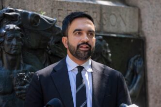 Zohran Mamdani, a man with a beard, speaks at a press briefing in a suit and tie in front of a stone monument.