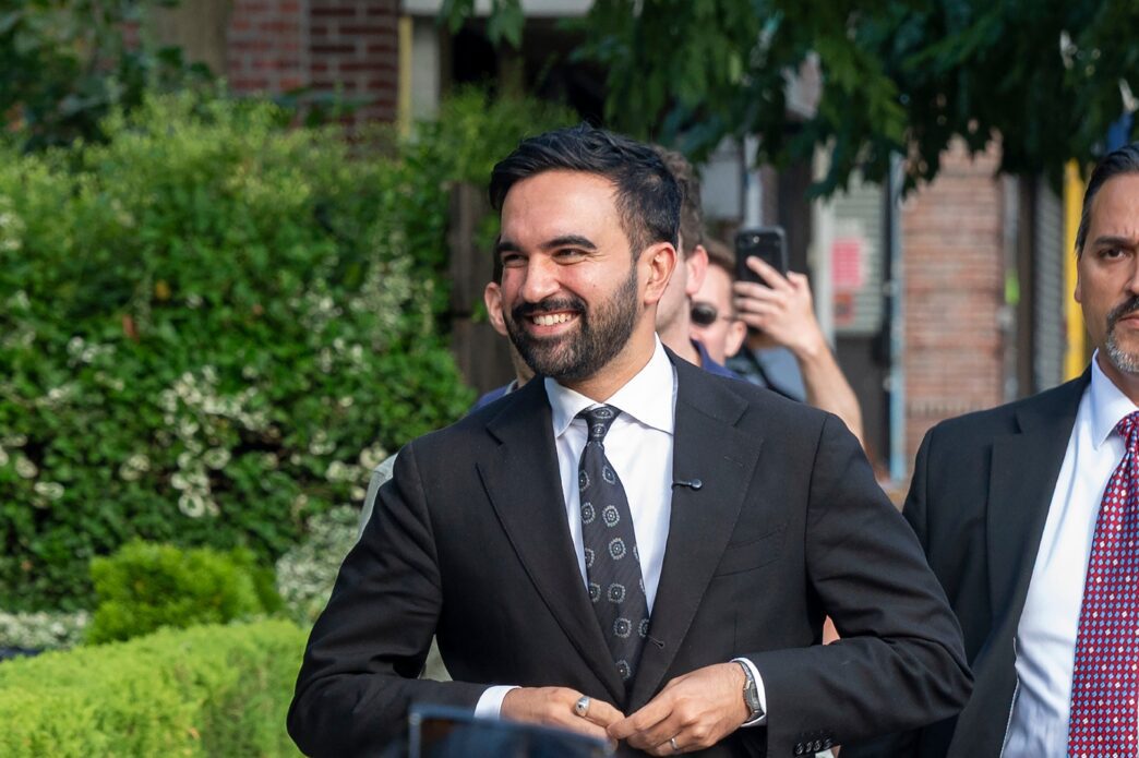 Democratic mayoral candidate Zohran Mamdani smiling outdoors in a black suit.