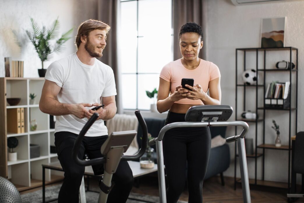 An African American woman in sportswear runs on a treadmill while a fit Caucasian man cycles on a stationary bike.