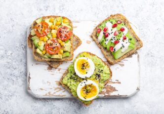 Overhead shot of avocado toast with egg, tomatoes, and seasonings on a white wooden cutting board.