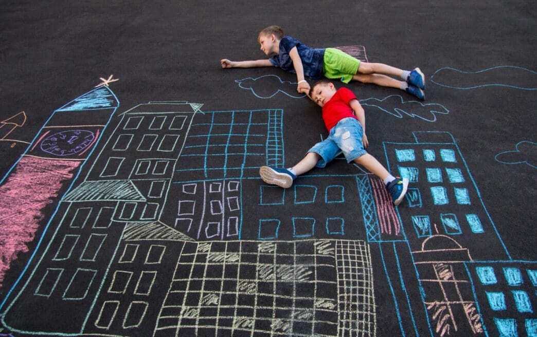 A chalk drawing on asphalt depicts a boy flying over a cityscape and saving a human superhero.