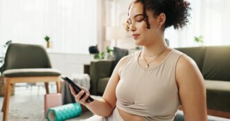 A woman in activewear uses a smartphone while practicing yoga at home.