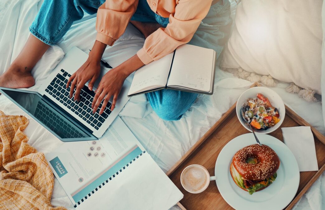 Woman in bed typing on a laptop with breakfast nearby, suggesting work from home and productivity.