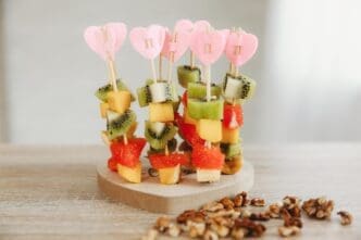 A heart-shaped stand displays colorful canapés of kiwi, mango, banana, and grapefruit on wooden skewers.