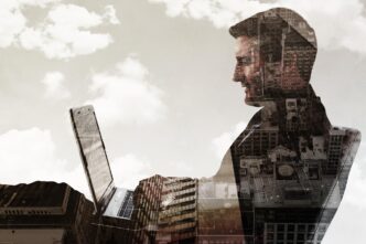 A businessman works on a laptop with skyscrapers visible in the background from a low-angle perspective.