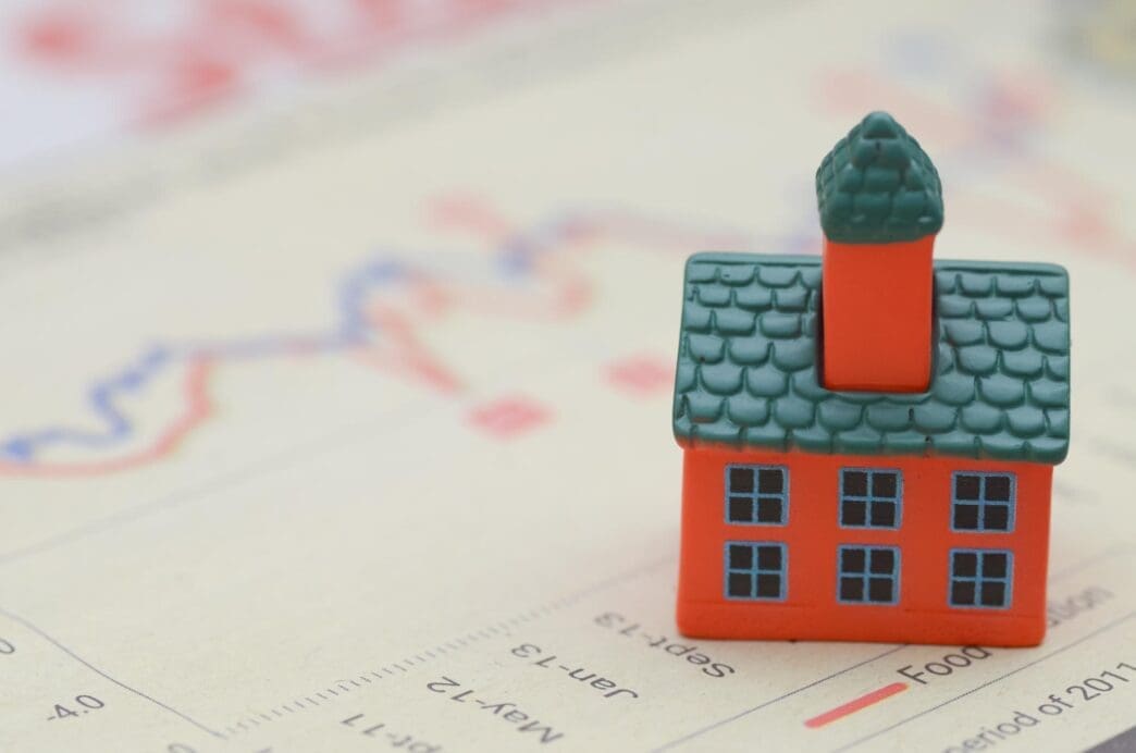 A miniature house sits atop a stack of newspaper headlines, illustrating the rising cost of living.