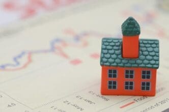 A miniature house sits atop a stack of newspaper headlines, illustrating the rising cost of living.