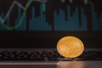A digital currency coin rests on a keyboard, with a stock chart visible in the background.