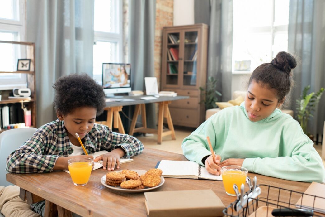 Children in a living room are taking notes in notebooks at a table, with a computer visible in the background.
