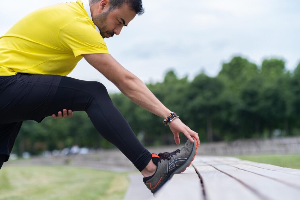 A man in black leggings stretches his leg in an urban park.