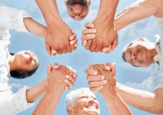 A family smiles and holds hands in a circle outdoors under a blue sky, demonstrating support and connection.