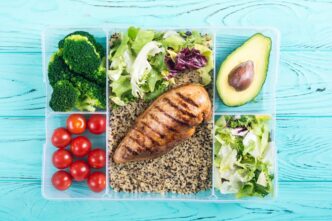 A clear plastic container holds a colorful and healthy lunch of quinoa, chicken, tomatoes, broccoli, avocado, and salad.