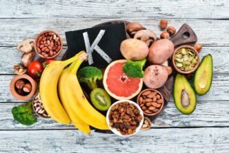 Overhead view of a white wooden surface displaying a variety of foods high in potassium, including potatoes, mushrooms, bananas, tomatoes, nuts, beans, broccoli, and avocados.