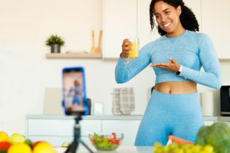 An African American woman films a video about healthy eating, holding a glass of juice and speaking to a phone camera.