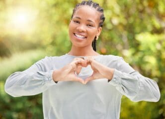 A Black woman forms heart hands, smiling outdoors in a park, possibly representing love, environmental care, or happiness.