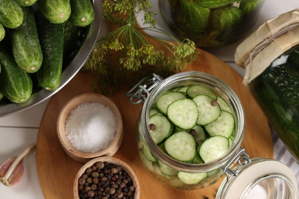A jar of homemade pickles with fresh cucumbers, dill, and spices sits on a white wooden table.