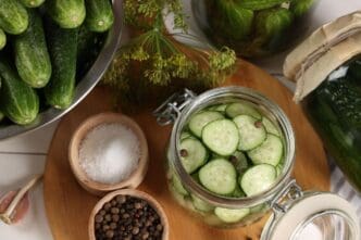 A jar of homemade pickles with fresh cucumbers, dill, and spices sits on a white wooden table.