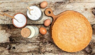 A top-down shot showcases a homemade sponge cake surrounded by ingredients like eggs and flour on a white wooden table.